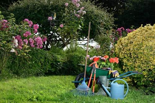 A well-maintained garden scene featuring a lush, green grass lawn in the foreground, bordered by vibrant flowering shrubs including pink and purple hydrangeas and a yellow-flowering bush. In the background, there are mature trees and dense foliage providing a natural enclosure. Garden tools such as a blue rake, a watering can, and a green wheelbarrow filled with gardening supplies are placed on the lawn, indicating ongoing gardening activity. The scene appears to be outdoors on a clear day, with natural lighting highlighting the various plant textures and colors, suggesting recent tending by a professional gardening service like Lawn Mowing Cricklewood in North West London. The layout shows a tidy, landscaped yard with a mix of ornamental and flowering plants, bordered by wooden stakes supporting some taller plants, creating a pleasant and inviting outdoor space suitable for maintenance and landscaping efforts.