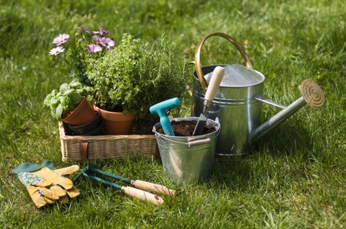 A neatly maintained front garden area with a vibrant green lawn featuring evenly cut grass. In the foreground, there are gardening tools including a pair of yellow work gloves, a trowel, and a small hand rake laid on the grass. To the left, a rectangular woven basket contains several potted plants with fresh green foliage and flowering plants exhibiting purple and white blossoms. Next to the basket, a metallic watering can and a bucket are placed, with the watering can having a long spout and a wooden handle. The background shows a slightly textured, lush grass surface extending beyond the immediate garden area, creating a natural outdoor space ideal for gardening and lawn care services. The scene suggests a well-organized outdoor space suitable for activities like planting, watering, and lawn maintenance in Cricklewood, reflecting professional gardening care. The lighting indicates a bright, possibly midday outdoor environment.