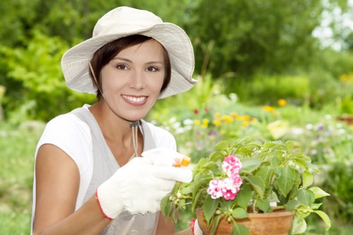 A woman wearing a wide-brimmed white hat, a white short-sleeved top, and gardening gloves is smiling while tending to a flowering plant in a terracotta pot outdoors. The background features a lush, green garden with various blooming flowers, shrubs, and a tree with dense foliage, indicating a well-maintained outdoor space typical of a residential garden in Cricklewood. The scene is brightly lit, suggesting a sunny day, and the woman appears to be engaged in gardening or plant care, which aligns with outdoor landscaping services offered by Lawn Mowing Cricklewood. The garden layout includes a mix of flower beds and green lawn areas, providing a natural and inviting environment suitable for lawn care and general garden maintenance.