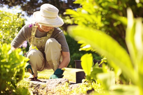 A woman wearing a wide-brimmed hat, glasses, and gardening attire is tending to a garden in a residential outdoor space. She is crouched down on a well-maintained lawn, surrounded by lush green plants, bushes, and foliage. The garden features a neatly trimmed grass area with a stone edging visible in the foreground, alongside a variety of leafy shrubs and flowering plants. In the background, there are tall trees providing shade, and the yard appears to be part of a landscaped front or back garden in Cricklewood. The scene is well-lit with natural sunlight, indicating a clear, bright day. The woman's activity involves planting or weeding, contributing to outdoor maintenance and garden care. This setting exemplifies typical garden landscaping and lawn care practices in a suburban UK environment, consistent with services offered by Lawn Mowing Cricklewood.