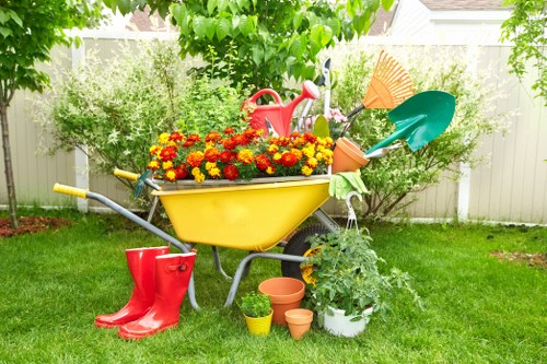 A garden scene featuring a yellow wheelbarrow filled with vibrant red, orange, and yellow flowering plants, positioned on a well-maintained lawn. Surrounding the wheelbarrow are gardening tools including a red watering can, a green spade, and a blue rake, with some smaller terracotta and plastic pots placed on the grass near the base. In the background, there are lush green shrubs and a hedge, with a white fence and part of a residential house visible. The scene appears to be outdoors on a bright, sunny day, suggesting active garden maintenance or planting in progress. This setting captures elements of garden care and landscaping, ideal for a professional gardening service website focused on outdoor lawn and garden improvements in Cricklewood.